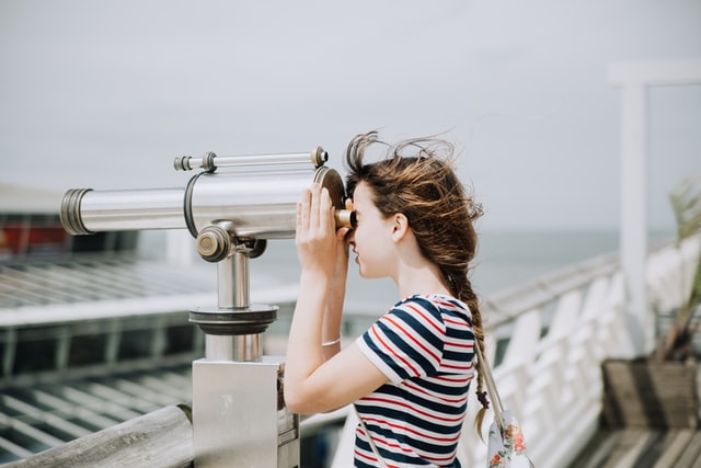 Fotografía de una chica mirando a través de un telescopio en un paseo marítimo