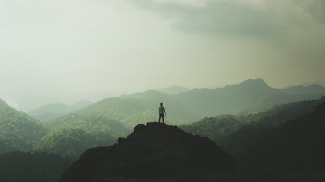 Fotografía de un hombre en la cima de una montaña rodeado de más montes y con niebla